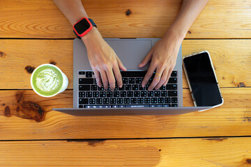 Close-up to  hands of a woman on  wrist, wearing a digital watch, typing on laptop keyboard placed on a wooden table, with a mobile phone and a glass of macha tea drink on the side