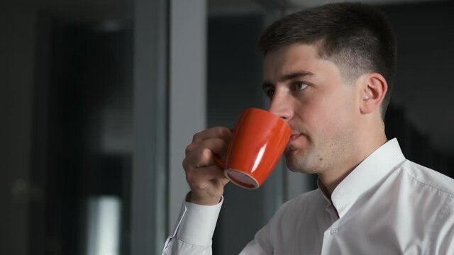 Handsome caucasian successful businessman man lingering at work and standing in evening office near panoramic window overlooking night city and drinking coffee or tea
