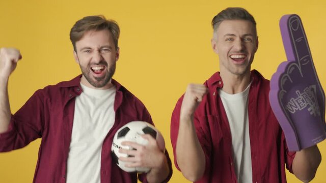 Two emotional football fans are screaming with football stuff standing isolated over yellow background