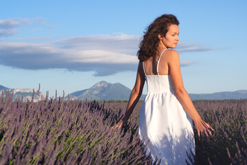 Summertime in lavender. Young beautiful woman in lavender flowers field at sunset in white dress....