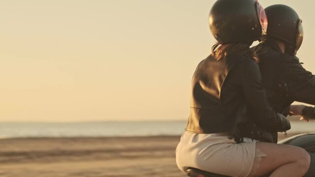 An attractive young couple man and woman are riding on a motorbike together on the beach during sunset time