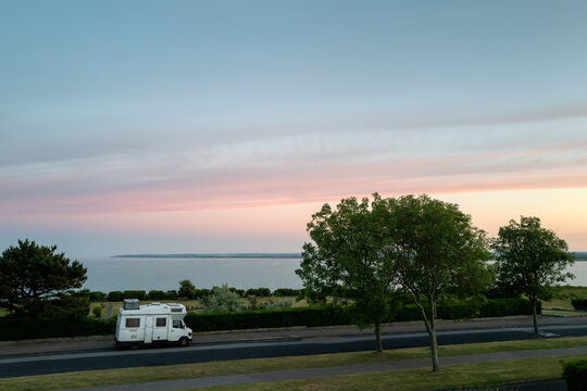 A vintage style camper van, motorhome with a metal trunk attached to its roof parked on a seafront road at sunset.