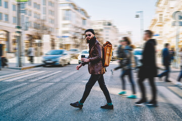 Full length portrait of male tourist with backpack from designer crossing across road and looking at camera during weekend in Russia, hipster guy in sunglasses walking and enjoying trip to Moscow