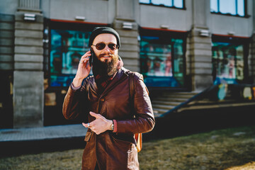 Fototapeta premium Happy smiling male tourist in stylish sunglasses enjoying positive smartphone conversation with friend using good internet connection in roaming during exploring streets in Moscow at autumn time