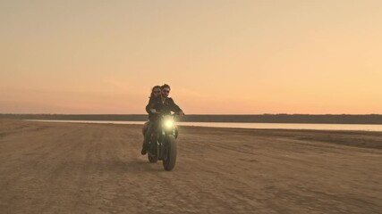 A stylish young couple man and woman are riding on a motorbike together on the beach during sunset time with headlight on