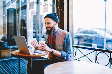 Happy hipster guy feeling good from received email from friend reading and recalls pleasent moment while using wireless 4g internet for communication on laptop computer during coffee break indoors