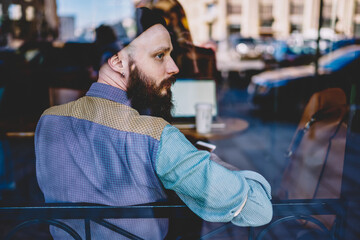 Thoughtful hipster guy in hat sitting near cafeteria window and thinking about life, pensive man...