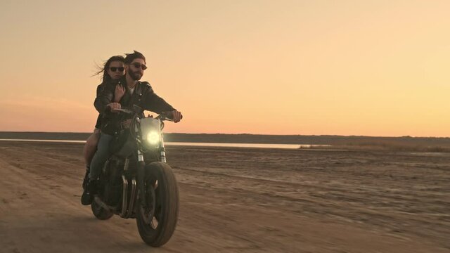 A happy attractive young couple man and woman wearing sunglasses are riding on a motorbike together on the beach during sunset time with headlight on