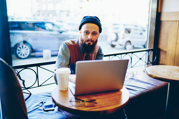 Caucasian hipster guy typing message to customer service for consultancy online and solving problems with internet connection, trendy man working remotely in coffee shop using laptop computer
