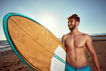 young caucasian beardy male walking on the sunny beach, holding surfboard, smiling. ultraviolet...