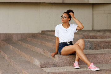 African girl in a white T-shirt and glasses sits on the steps. Summer