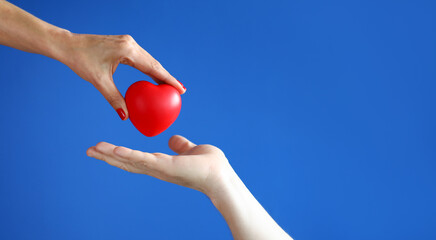 Female hand passes red heart to male hand against blue background closeup. Kindness and charity concept