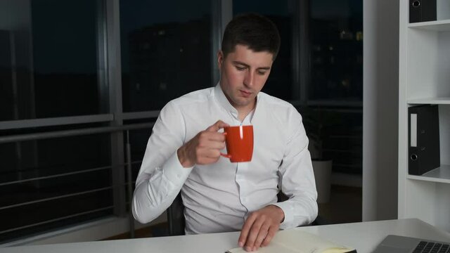 An office worker with a serious face lingers at work until late at night sitting at his workplace in an office with panoramic windows and working. Makes a business plan for the team for tomorrow