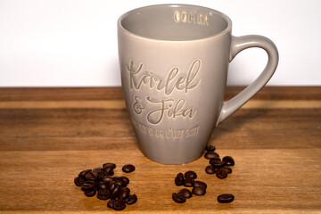 Coffee cup says "Love and Fika" on a wooden table with coffebeans infront on a white background