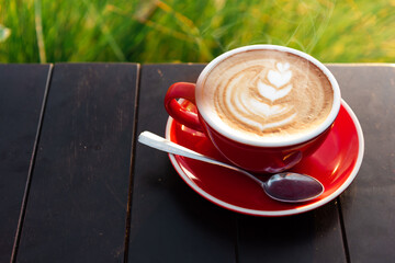 Red cup of coffee placed on wooden desk in front of green field.