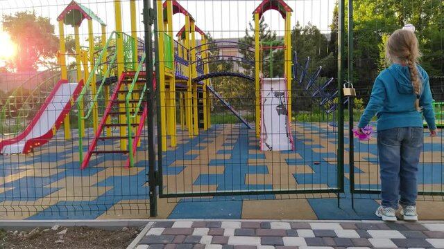 Sad lonely child near the closed playground because of the quarantine.A girl with long blonde braided hair staying alone near empty fenced showground without children.COVID-19 lockdown social distance