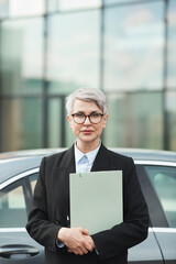 Fototapeta premium Portrait of mature businesswoman in suit and in eyeglasses holding documents and looking at camera while standing in the city