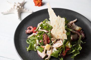 Octopus salad with tomato and rocket in a black plate on rustic wooden background.