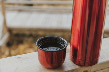 Red thermos bottle with cup in wooden house on background of wwoden trail. Close up.