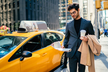 Handsome male in elegant suit walking on Manhattan near yellow cab reading information from daily...