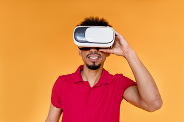 Close up portrait of young afro american man posing over isolated orange background wearing summer clothes and wearing virtual reality headset or 3d glasses