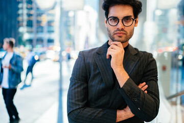 Half length portrait of male owner of business in elegant suit and spectacles looking at camera on urban setting, handsome proud ceo dressed in formal wear standing with crossed arms on city street.