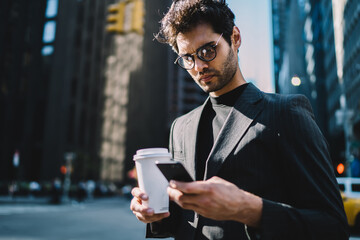 Confident male owner in elegant formal wear using online app for calling taxi standing on avenue with coffee to go, serious businessman checking mail on telephone and sending feedback on street.
