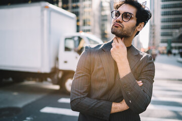 Contemplative male entrepreneur in trendy formal clothing thinking about business idea on Manhattan, handsome pondering executive in fashion suit standing with crossed arms on avenue in downtown.