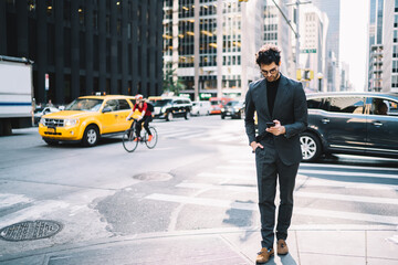 Successful businessman dressed in formal wear strolling on Manhattan street and reading financial news in app on smartphone.Executive director in elegant suit walking in urban setting with cellular