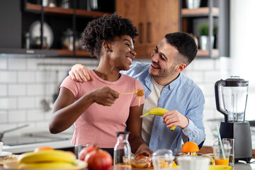 Fototapeta na wymiar Multiethnic couple preparing healthy smoothie with tropical fruits and smile in kitchen