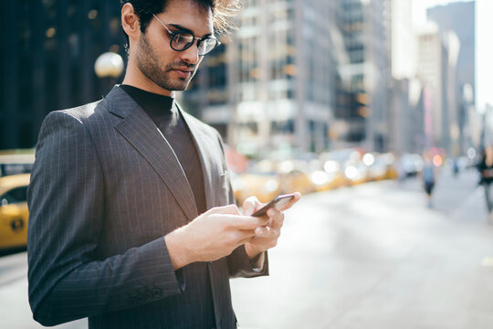 Cropped image of confident executive manager in elegant suit reading last business news received on mail via smartphone.Serious entrepreneur sending message on telephone standing on New York avenue
