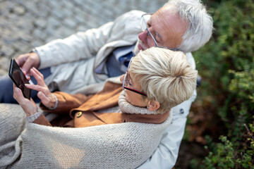 Mature couple sitting on bench in public park and looking at mobile phone