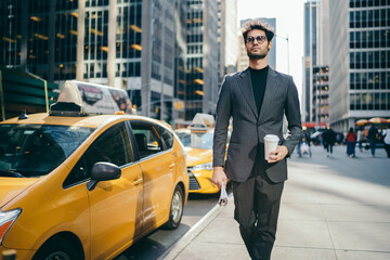 Serious male manager 30s dressed in elegant trendy wear and eyeglasses spending time outdoors, handsome businessman with coffee to go walking near traffic road with taxi in metropolitan downtown