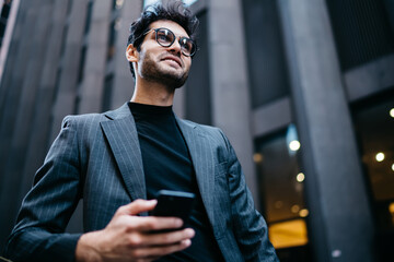 Young positive male in formal suit and spectacles walking on street using mobile phone and 4G...