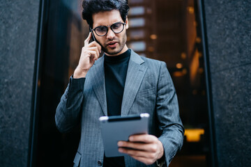 Young businessman furrowing brows while talking via cellphone and looking at information on tablet