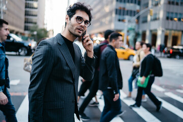 Prosperous young male in trendy stylish suit and spectacles having mobile phone conversation strolling on noisy street , smiling successful businessman using cellular walking on pedestrian crossing.