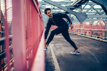 Fototapeta na wymiar Handsome european sportsman in trendy tracksuit stretching on city bridge before start running across street, young jogger doing workout outdoors during listening radio via electronic headphones