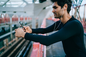 Side view of sportsman dressed in active wear for workout listening music in earphones connected to smartphone holding in hands.Fit man choosing audio app for installing on phone for sport training