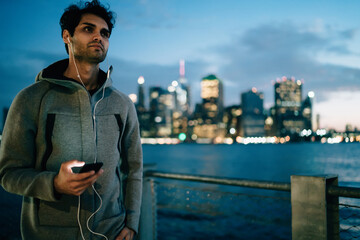 European male runner listening carefully guides for evening jogging using mobile application and electronic headphones while standing on blurred background with city, concept of healthy lifestyle
