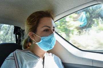 A young passenger woman wearing a respiratory protective face mask looks at the landscape in the back of a vehicle during a travel. Concept of "new normality"