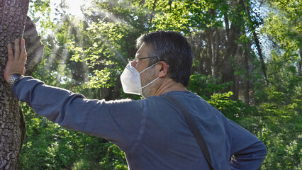 A hiker wearing face mask enjoys the sun rays filtering through the trees branches while resting on a walk during the coronavirus pandemic