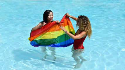 Two women of different race play with a rainbow flag inside a pool. Female friends or lgbt couple celebrating lgbt pride day.