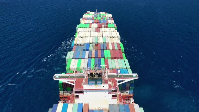 Aerial drone top down tracking video of huge Container cargo Ship carrying load in truck-size colourful containers cruising deep blue Aegean sea, Greece