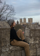 Photo of a young and attractive woman sitting on a wall in York wearing winter cloths and a grey hat. 