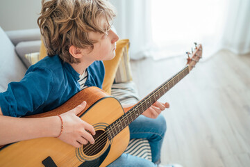 Top angle view at Preteen boy playing acoustic guitar dressed casual jeans, blue shirt sitting on the cozy sofa at the home living room and thinking about something. Music education concept image.