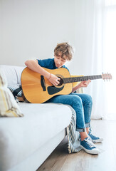 Preteen boy playing acoustic guitar dressed casual jeans, shirt and new sneakers sitting on the cozy sofa at home living room. Music education top angle view concept image.