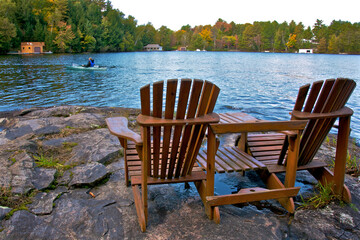 Naklejka premium Retirement Living - Two Muskoka chairs sitting on a rocky shore facing a calm lake with cottages in the background