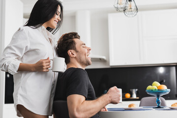 Side view of sexy woman in shirt holding cup of coffee near cheerful boyfriend in kitchen