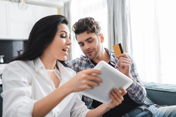 Selective focus of man holding credit card near excited girlfriend showing digital tablet at home