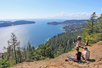 Soames Hill park in Sunshine Coast, BC. The view on the 2 couple sitting and resting on the hill with the aerial view on Gibsons surrounded by forest / woods and ocean.  - obrazy, fototapety, plakaty